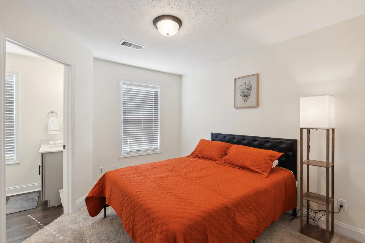 A cozy bedroom featuring a vibrant orange quilted bedspread atop a black upholstered headboard, flanked by a modern floor lamp and framed artwork, offering a warm, inviting atmosphere with soft lighting ideal for relaxation.