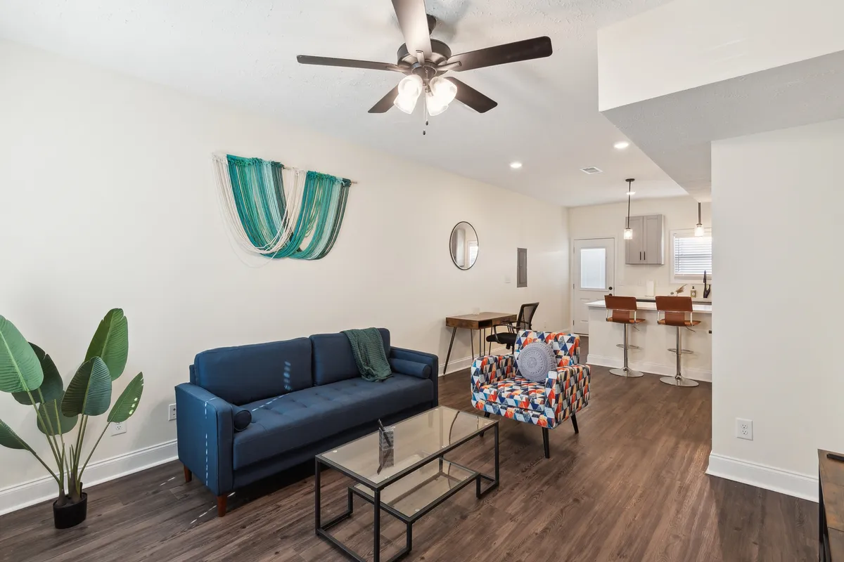 The image showcases an inviting living space featuring a plush blue sofa and a vibrant accent chair, both arranged around a sleek glass coffee table. The open layout flows seamlessly into a modern kitchen, suggesting an ideal setting for family gatherings, relaxing evenings, or casual meals. Warm lighting and tasteful decor enhance the comfortable and welcoming atmosphere.