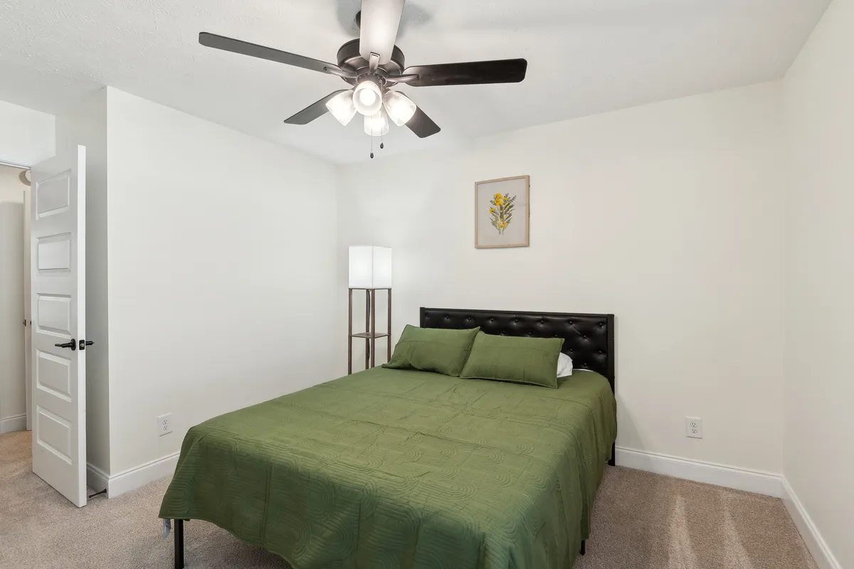 A bright, serene bedroom is shown with green bedding, a ceiling fan, bedside lamp, and framed artwork. The atmosphere is peaceful, with soft lighting inviting moments of rest or quiet reading.