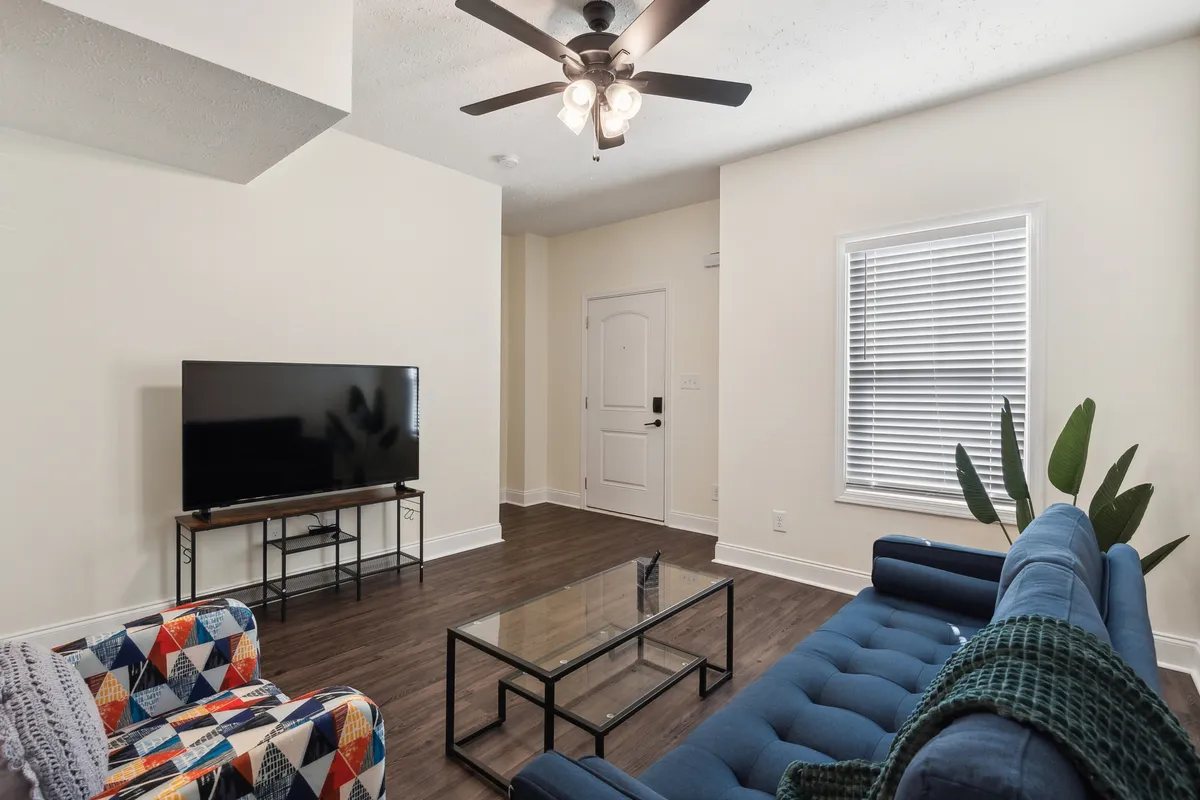 This image presents an inviting living room scene with a blue sofa and a colorful armchair, oriented around a glass coffee table. The television adds to the entertainment possibilities, while the lighting and pops of color create a cozy, home-like ambiance.