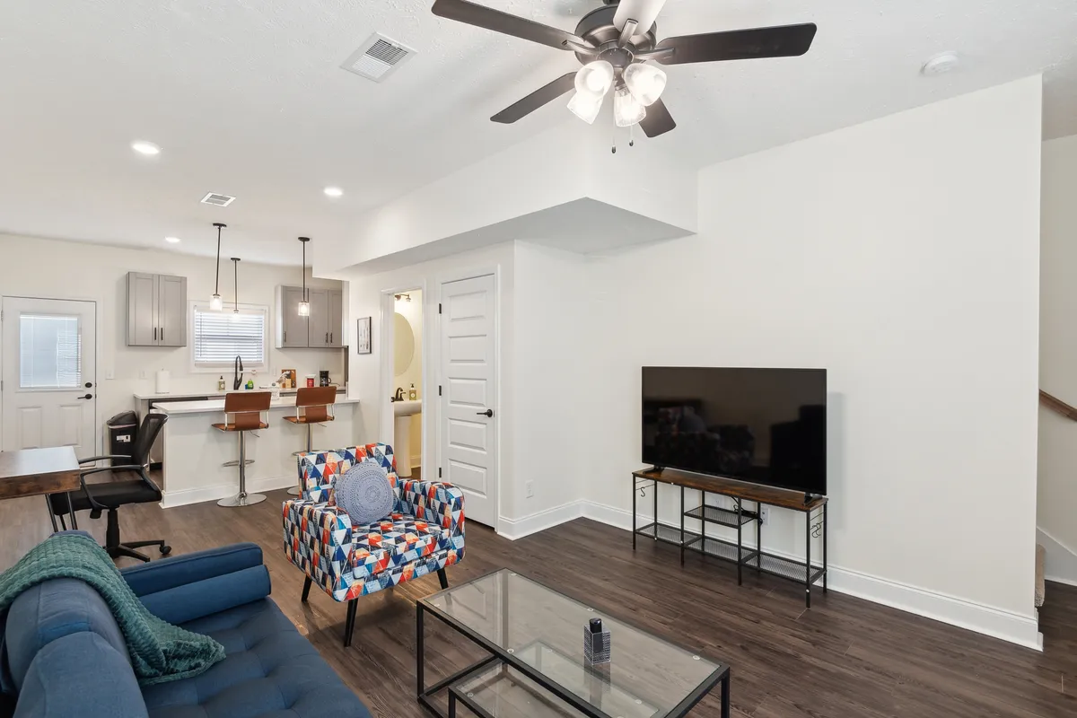 The image features a colorful armchair and a modern sofa arranged in an open-plan living area. A glass coffee table sits centrally, and a television completes the comfortable entertainment setup. The space is bathed in soft lighting, with accents of vibrant decor and a practical layout that promotes relaxation and socializing.